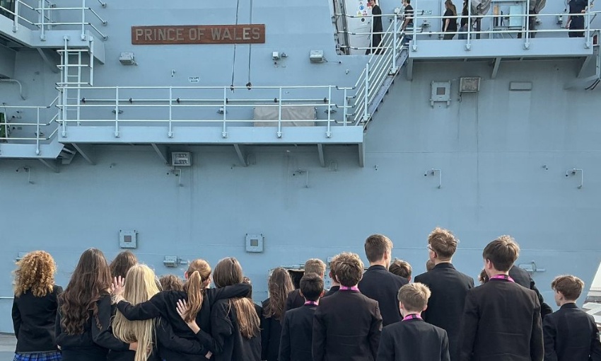 Pupils in uniform look up at a ‘PRINCE OF WALES’ sign mounted on the side of a grey naval ship.